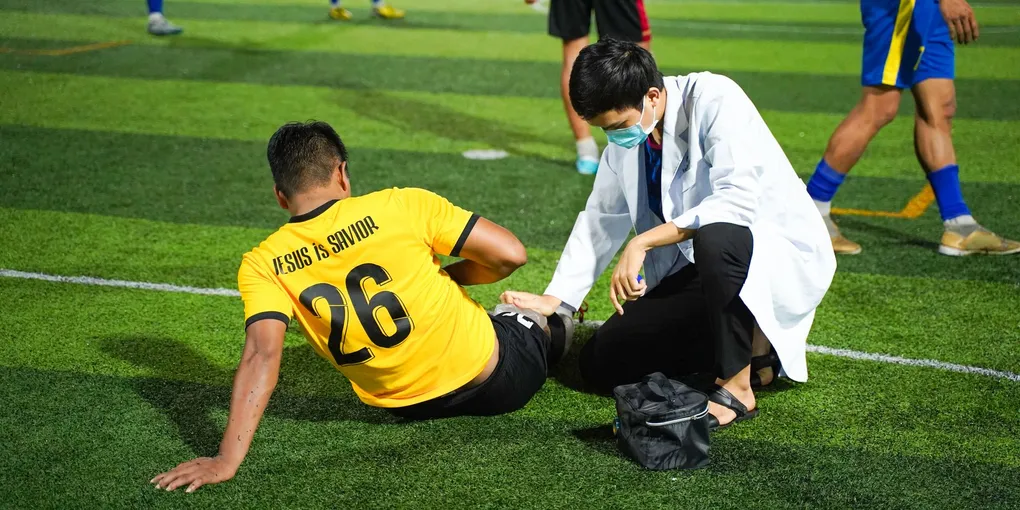 Atleta sentado no gramado segurando a parte posterior da coxa após uma lesão